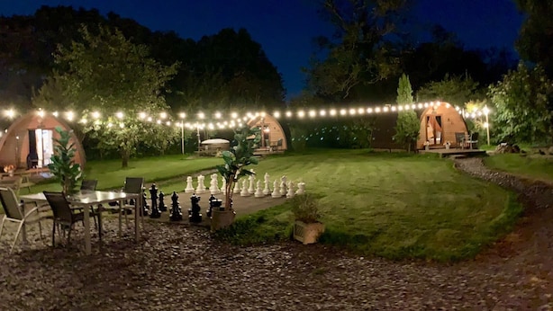 A nighttime outdoor setting with two wooden dome structures, string lights, and a chessboard arrangement. - Dog Friendly Cozy Bell Tent With Log Burner (Gaerwen)