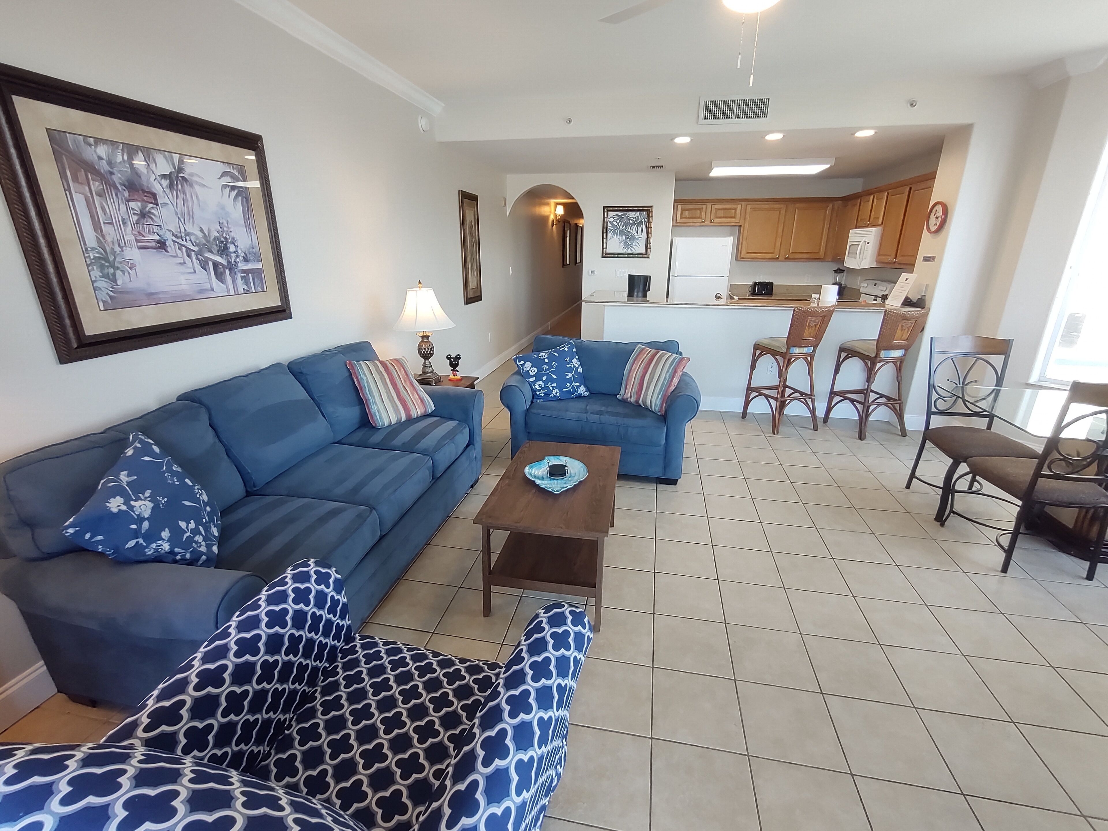 A living room with a blue sofa, a patterned armchair, and a coffee table. There is a kitchen area with wooden cabinets and a refrigerator.