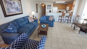 A living room with a blue sofa, a patterned armchair, and a coffee table. There is a kitchen area with wooden cabinets and a refrigerator.
