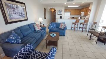 A living room with a blue sofa, a patterned armchair, and a coffee table. There is a kitchen area with wooden cabinets and a refrigerator.