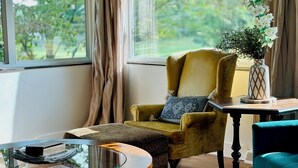 Una habitación con un sillón amarillo, una mesa de centro redonda de cristal, una mesita auxiliar con un jarrón y vistas a zonas verdes.