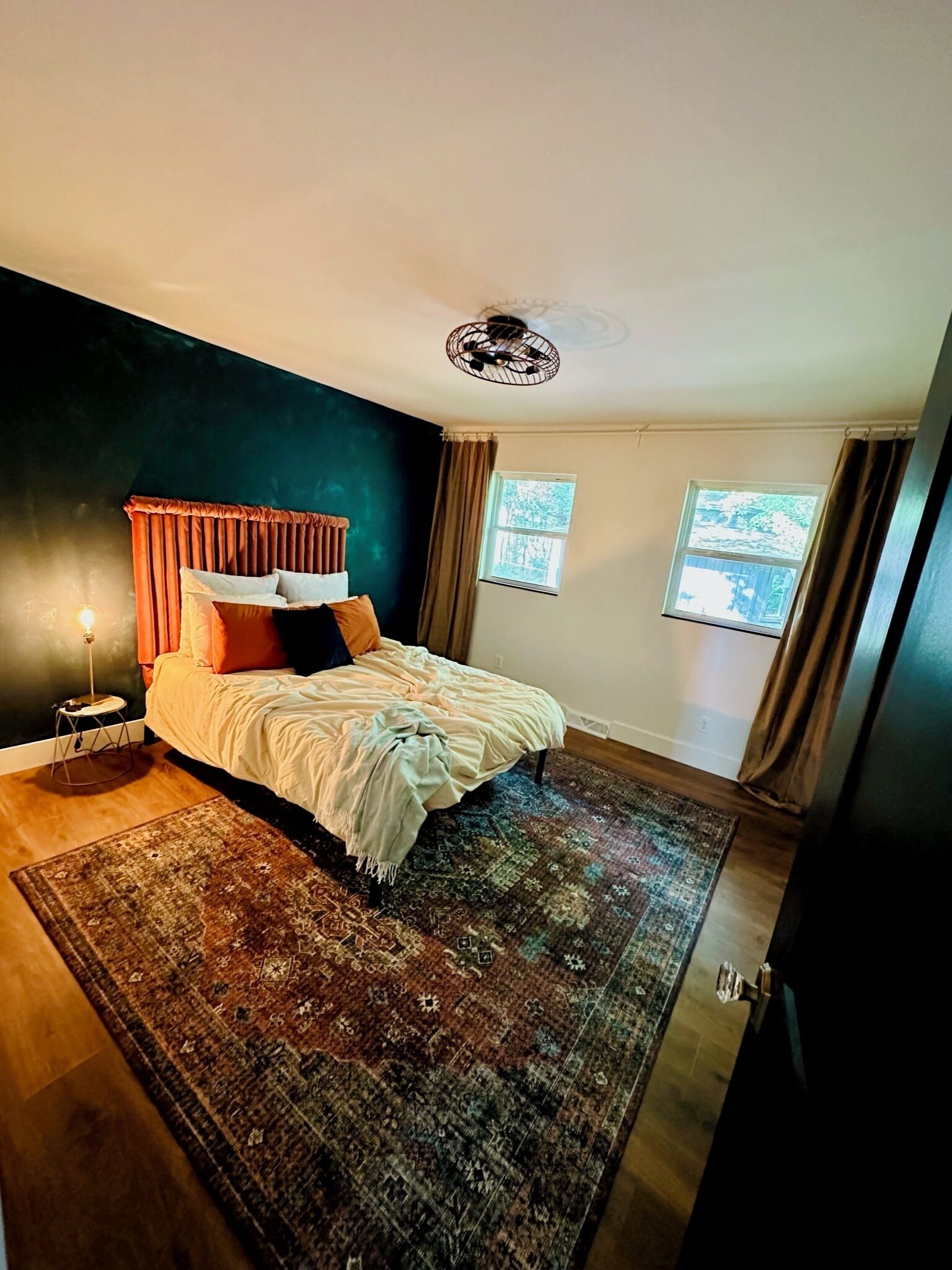 Une chambre à coucher comprenant un lit, une table de chevet, une lampe sur pied, un tapis à motifs et deux fenêtres avec des rideaux.
