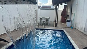 A swimming pool with a water feature, a patio area with a table and chairs, and a brick structure in the background.
