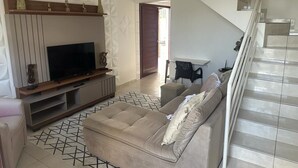 A modern living room with a flat-screen TV, a beige sofa, and a staircase.