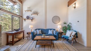 A cozy living room with a blue sofa, wooden coffee table, and a large window offering a view of trees. - 2 BR/2 BA Condo by Northstar (Truckee)