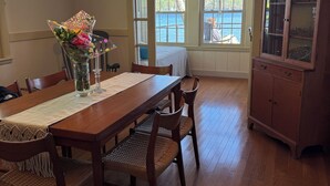 A dining room with a wooden table, chairs, a chandelier, and a view of the outdoors through large windows. - Charming 4-bedroom cottage with AC, WiFi on Little Sebago Lake waterfront (Raymond)