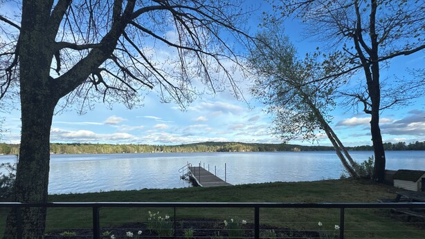 A lakeside view from a deck with a dock extending into the water, surrounded by trees and a grassy area with flowers. - Charming 4-bedroom cottage with AC, WiFi on Little Sebago Lake waterfront (Raymond)