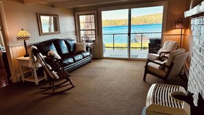 A cozy living room with a leather sofa, rocking chair, and armchairs, a fireplace, and a view of a lake through large windows. - Charming 4-bedroom cottage with AC, WiFi on Little Sebago Lake waterfront (Raymond)