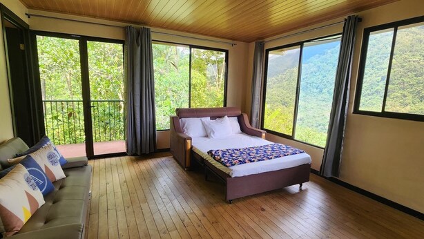 A bedroom with a bed, a sofa, and large windows offering a view of a forested area.