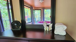 A room with a wooden ceiling, a window with a view of greenery, a fan on a table, and a stack of towels.