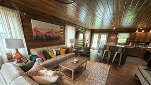 A cozy cabin interior with a wooden ceiling, a sofa with colorful cushions, a dining area, and a kitchen. - Luxury Cabin Hot Tub ATV Trail Cherry Springs (Coudersport)