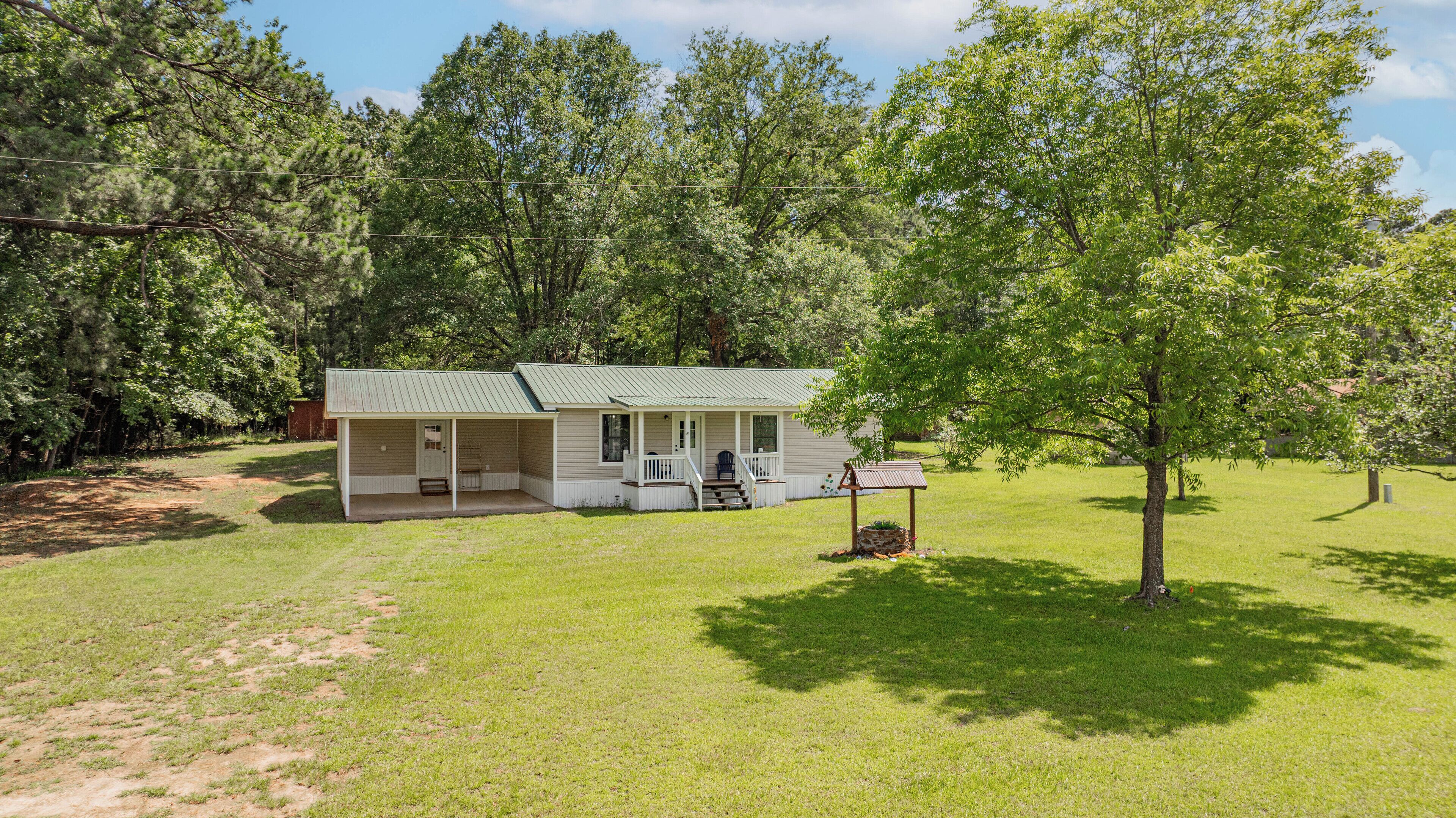 A single-story house with a porch and a covered outdoor seating area, surrounded by a grassy yard and trees.