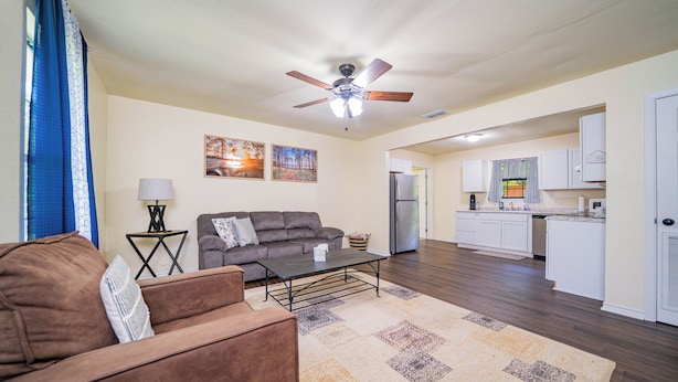A living room with a brown sofa, a coffee table, and a ceiling fan. There is a kitchen in the background with white cabinets and a refrigerator.