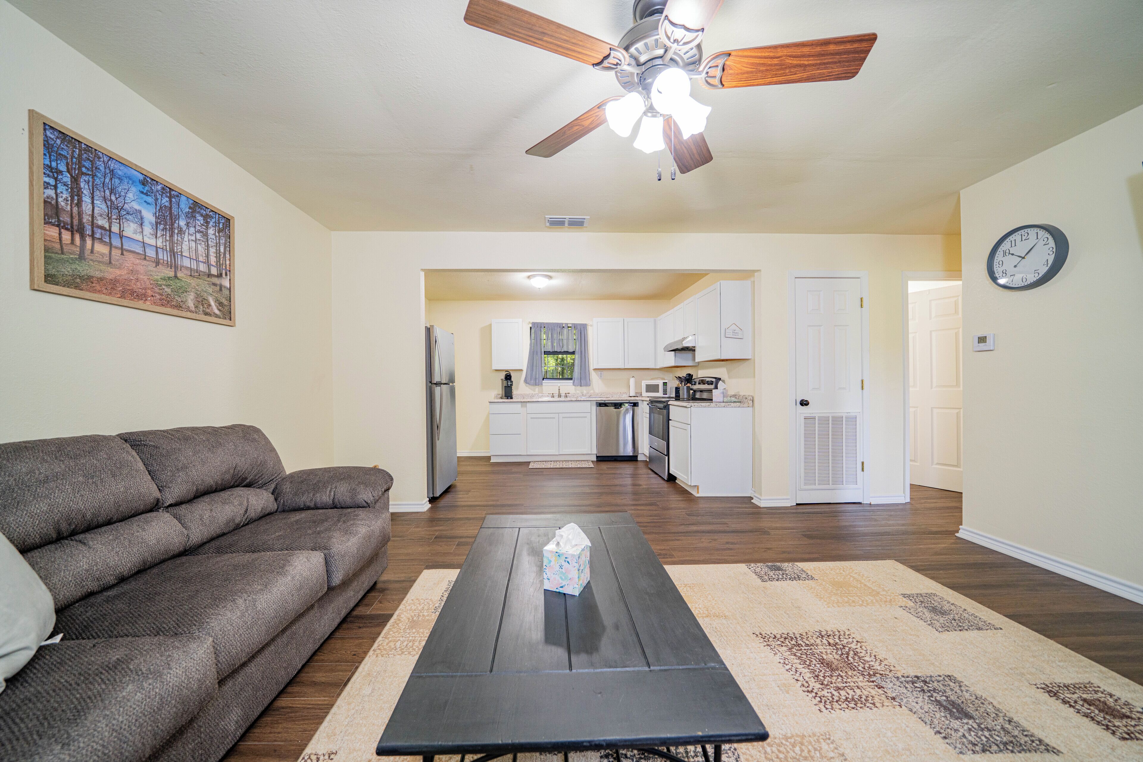 A living room with a sofa, a coffee table, and a ceiling fan. There is a kitchen visible through an open doorway.