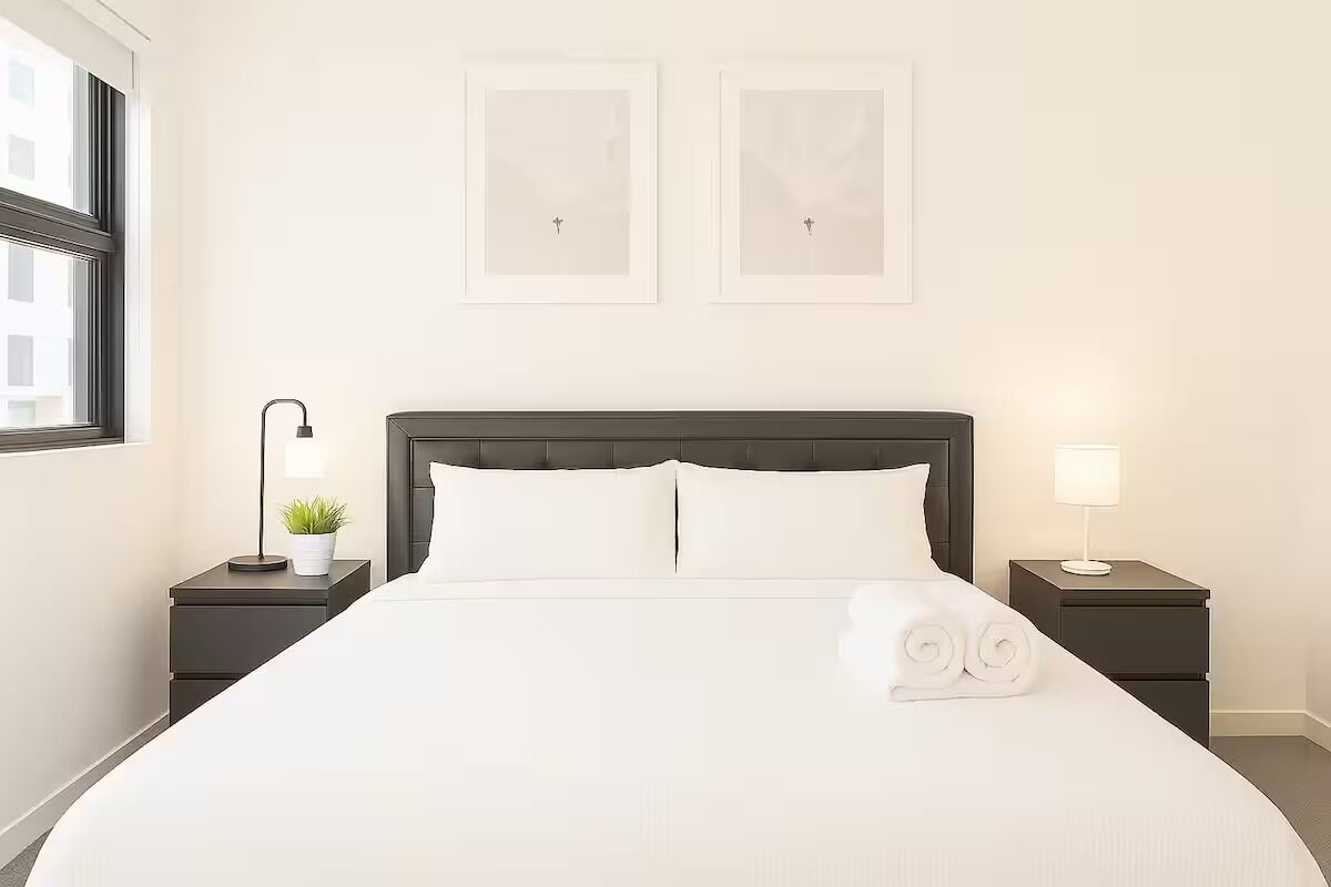 Una cama bien hecha con sábanas blancas y toallas enrolladas, dos mesitas de noche con lámparas, una pequeña planta en maceta y dos cuadros en la pared.