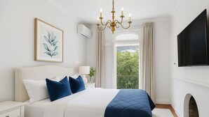 A bedroom with a large bed, a chandelier, a flat-screen TV, and a view of greenery outside. - The Hamptons Paddington Home (Paddington)