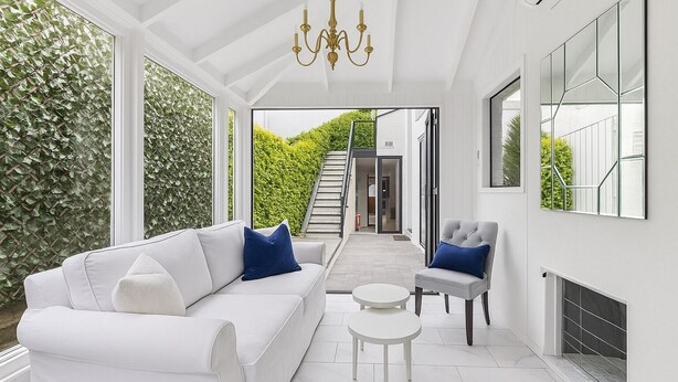 A modern living room with a white sofa, two blue cushions, a grey armchair, a small white table, and a large window with a view of a green garden and a staircase. - The Hamptons Paddington Home (Paddington)