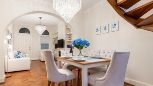 A dining area with a wooden table, white upholstered chairs, and a chandelier. - The Hamptons Paddington Home (Paddington)