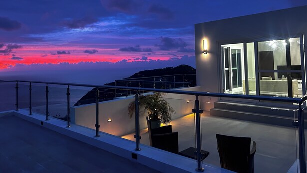 Un edificio moderno con fachada de vidrio, zona de estar al aire libre y vista al atardecer sobre el agua.