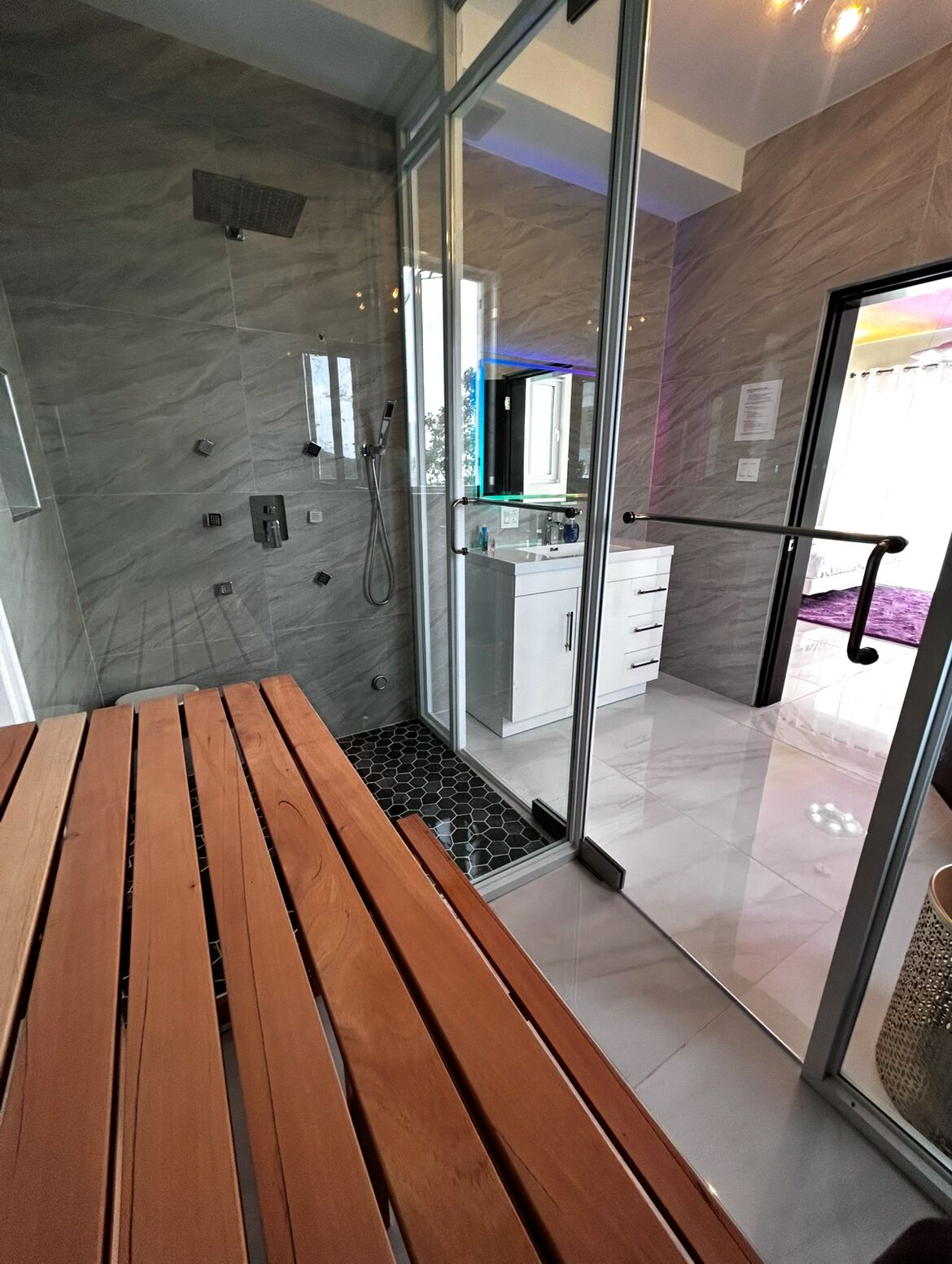 Un baño moderno con cabina de ducha de vidrio, un banco de madera y una cocina visible a través de las puertas de cristal.