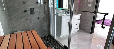 Un baño moderno con cabina de ducha de vidrio, un banco de madera y una cocina visible a través de las puertas de cristal.