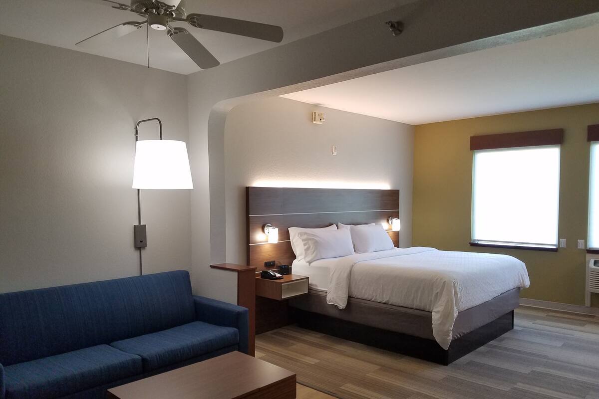 Quarto de hotel com cama, sofá azul, mesa de centro de madeira, ventoinha no teto, abajur fixado na parede e janela com persianas.