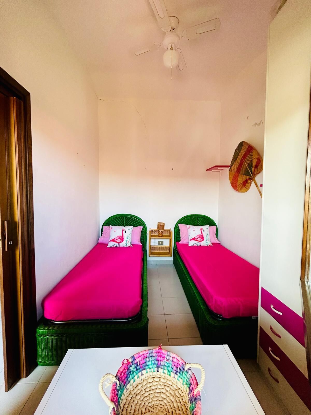 Quarto pequeno e limpo com duas camas de solteiro, um ventilador de teto, um ventilador de parede e uma pequena mesa com uma cesta de vime.