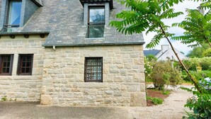 Un edificio de piedra con techo inclinado, una ventana con celosía decorativa y un jardín con un sendero.
