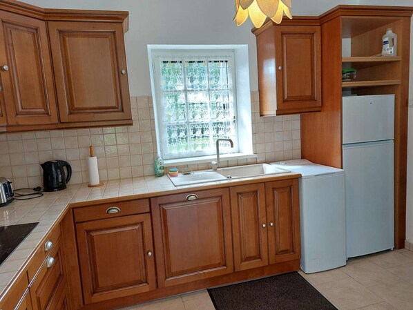Una cocina con armarios de madera, un refrigerador blanco, una ventana con cortinas de encaje y una tetera sobre la encimera.