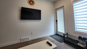 Ein modernes Wohnzimmer mit einem an der Wand montierten Flachbildfernseher, einem Holztisch, einer Fernbedienung, einem Schuhregal und einer Tür mit Jalousien.