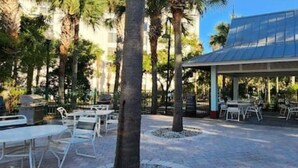An outdoor patio area with white tables and chairs, surrounded by palm trees and a covered seating area.