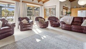 A spacious living room with leather sofas and armchairs, large windows offering a view of the outdoors, and a kitchen area with wooden cabinets.