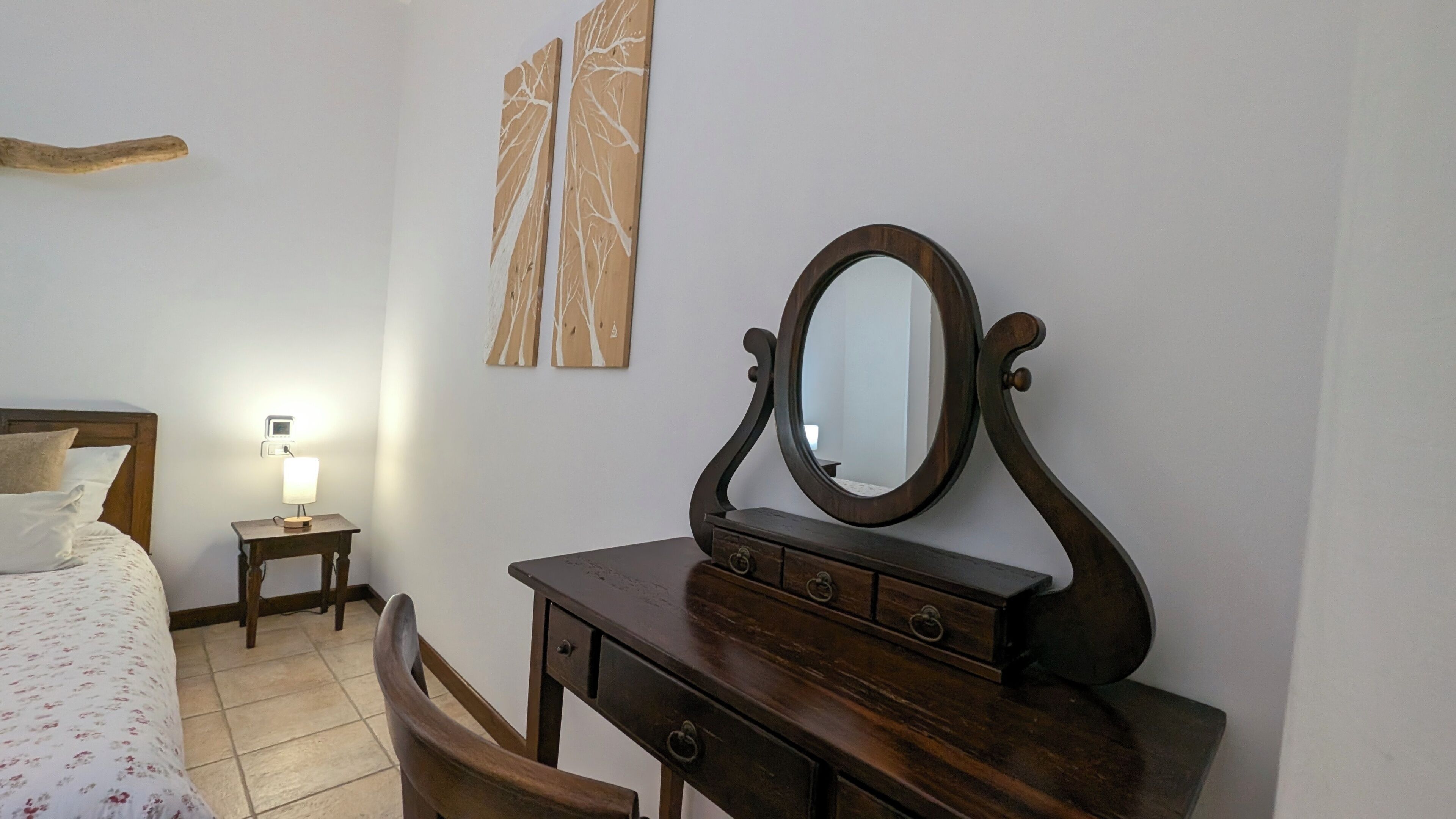 A bedroom with a wooden dresser, a mirror, a bed with a floral bedspread, a nightstand with a lamp, and two wall art pieces.