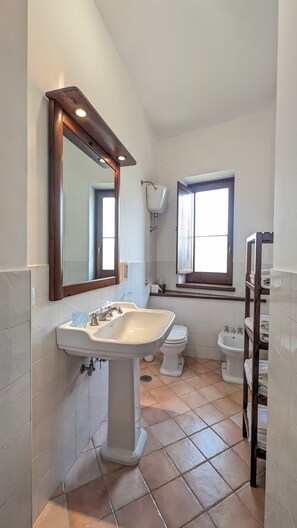 Un baño con lavamanos pedestal, dos inodoros, una ventana y un estante de madera.