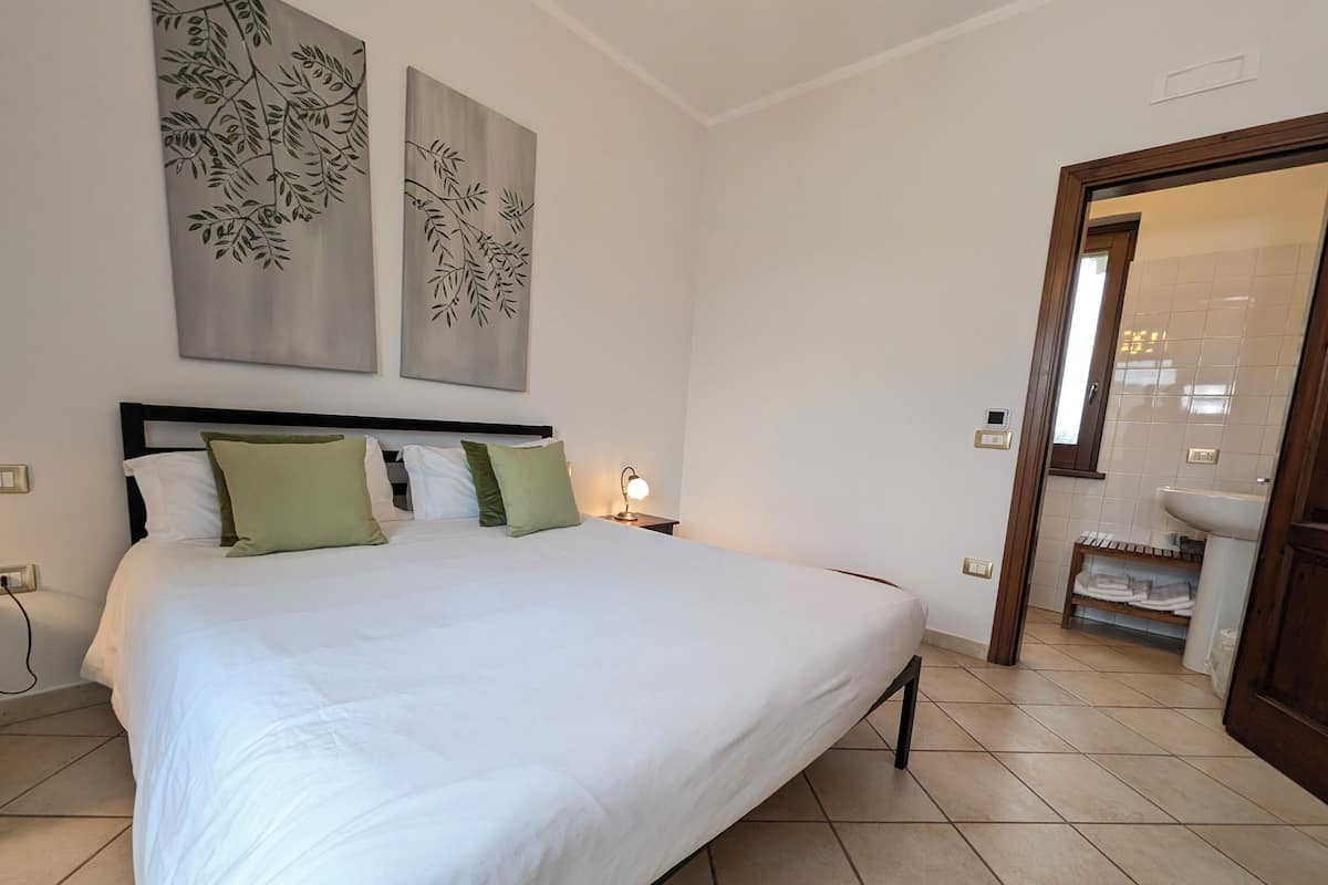 Una cama bien hecha con almohadas blancas y verdes, una mesita de noche con lámpara, y un baño visible a través de una puerta abierta.