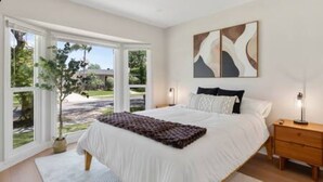 A bedroom with a large bed, a nightstand, and a window with a view of greenery. - Elegant Oasis | Heated Pool & Spa | 5mi to Beach (Long Beach)