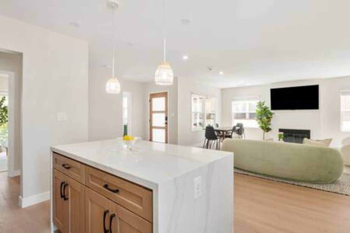 Una cocina moderna con una isla blanca, armarios de madera, iluminación colgante y un rincón de comedor con un sofá verde y un televisor de pantalla plana.