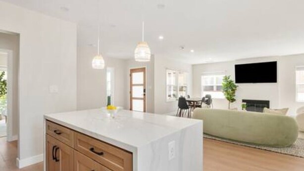 A modern kitchen with a white island, wooden cabinets, pendant lighting, and a dining area with a green sofa and a flat-screen TV. - Elegant Oasis | Heated Pool & Spa | 5mi to Beach (Long Beach)
