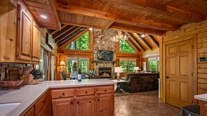 A log cabin interior with a stone fireplace, wooden cabinets, and a large window overlooking greenery. - *Riverhouse on the Cartecay*WaterFront~Tube~Family (Ellijay)