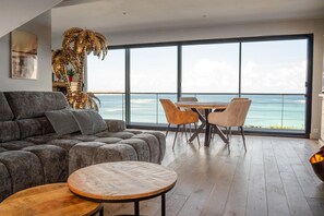 Un salon moderne doté d’une grande fenêtre donnant sur la mer, d’un canapé, d’une table ronde en bois et d’un coin repas avec des chaises.