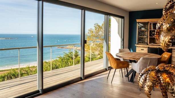 Habitación moderna con ventanas amplias que dan a una playa, una mesa de madera y sillas.