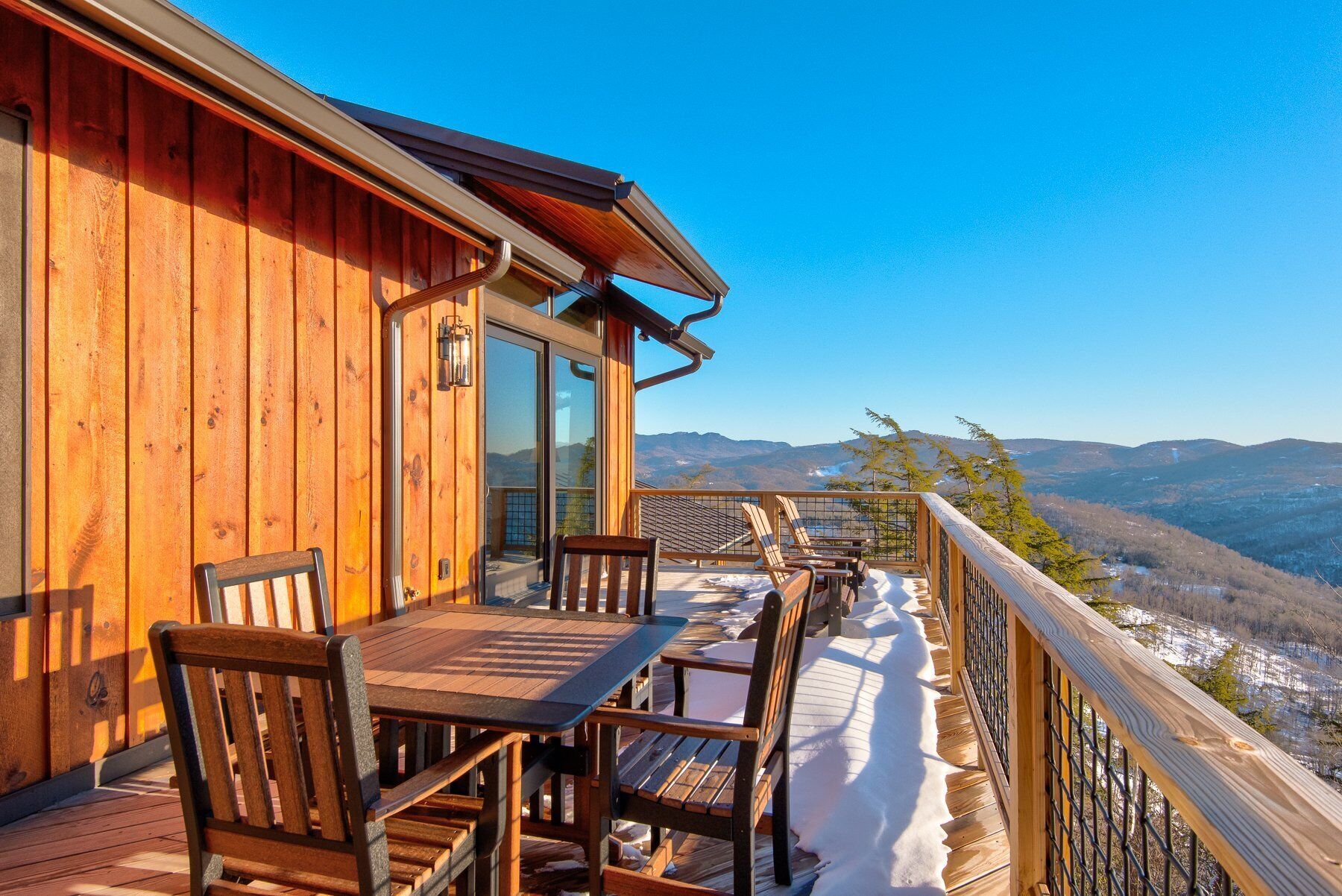 A wooden deck with a table and chairs, overlooking a snowy landscape and distant mountains.