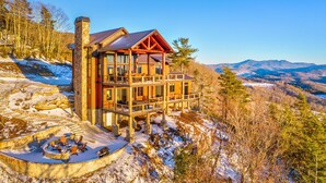 A multi-level wooden cabin with a stone chimney, surrounded by snow-covered terrain and forested mountains.