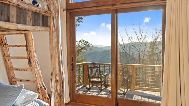 A room with a wooden ladder, a bed with pillows, and a large sliding glass door leading to an outdoor balcony with a chair.