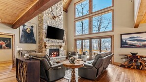 A spacious living room with a stone fireplace, leather sofas, and large windows offering a view of the outdoors. - Rock Haven at Eagles Nest - pets, views, four king beds, hot tub (Banner Elk)