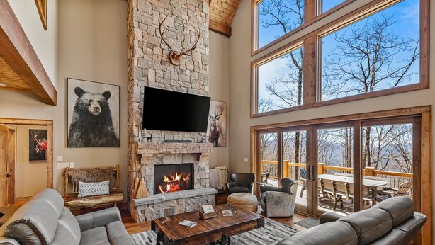 A spacious living room with a stone fireplace, leather furniture, and large windows offering a view of the outdoors. - Rock Haven at Eagles Nest - pets, views, four king beds, hot tub (Banner Elk)