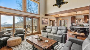 A spacious living area with a large window overlooking a deck, a fireplace, and a kitchen area with bar seating. - Rock Haven at Eagles Nest - pets, views, four king beds, hot tub (Banner Elk)