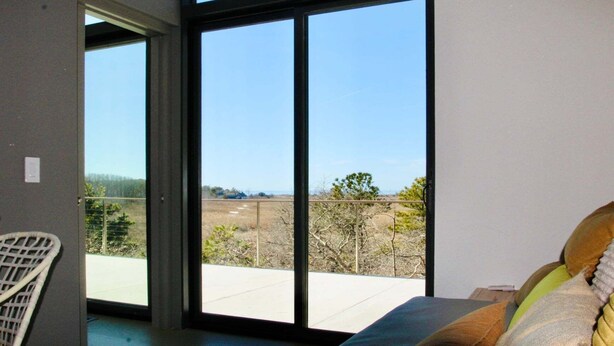 A room with a large sliding glass door leading to a balcony with a view of a rural landscape. - Cape Cod Bay Views | 2BR/2BA Eastham, Modern, EV (Eastham)