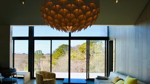 A modern living room with a large window, a sofa, a chair, and a unique ceiling light fixture. - Cape Cod Bay Views | 2BR/2BA Eastham, Modern, EV (Eastham)