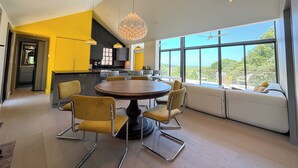 A modern kitchen with a central island, yellow chairs around a round table, and a large window offering a view of greenery. - Cape Cod Bay Views | 2BR/2BA Eastham, Modern, EV (Eastham)
